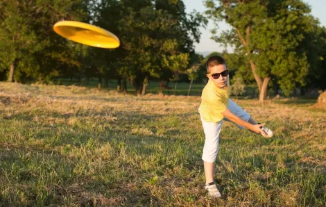 A school-age boy in a yellow shirt and sunglasses throws a yellow Frisbee disc during a game of disc golf free fun family sport to play in parks around Seattle