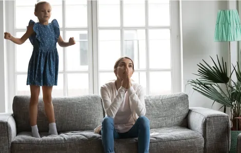 Frustrated mother sitting on couch while daughter jumps on the couch