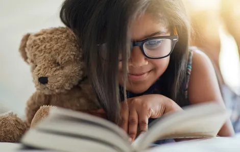 Girl cuddling stuffed bear, smiling and reading a book series for kids