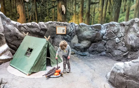 Young boy with curly hair tends a play campfire near a play camping tent on "the mountain" exhibit at Seattle Children's Museum reopening after a two-plus-year pandemic closure