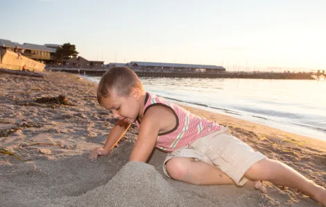 A young boy in shorts and a tank top plays in the sand on the beach in Edmonds, Washington. It's a sunny evening and a pier is visible behind him.