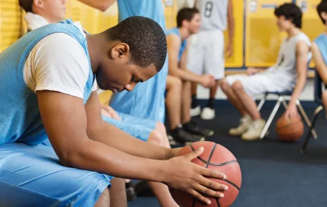 Male teenage basketball player holding a basketball in a locker room and looking at the floor