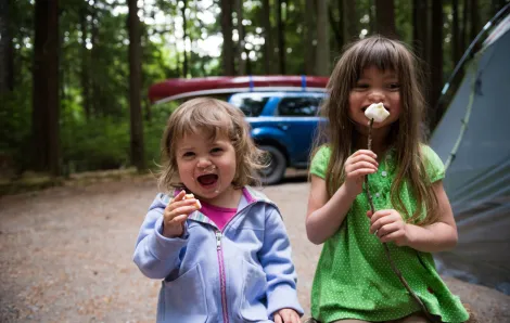 Two girls at a campsite; they're holding marshmallows on sticks and their family tent and car are visible in the background