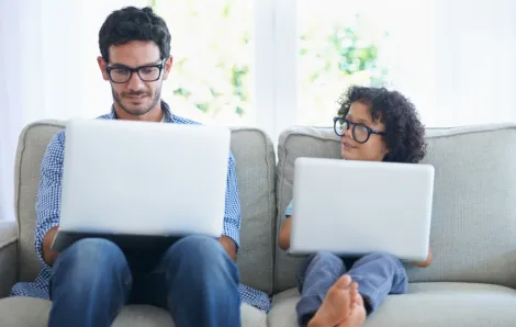 Man and child sitting on a couch each holding a laptop