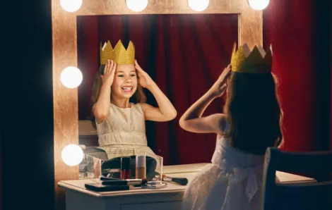 Young girl wearing a play crown sitting in front of a mirror with lights around it