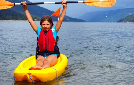 A girl in a yellow sit-upon kayak holds her paddle over her head in Shuswap Lake, British Columbia, among great lake vacations for summer for Seattle-area families