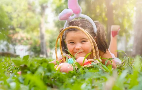 A cute young girl peers through the rounded handle of her Easter basket with colored eggs in it. She is lying in the grass and wearing bunny ears. Best Seattle Easter events for kids and families 2023