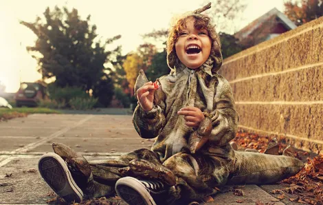 Little boy sitting on the sidewalk smiling wearing a dinosaur Halloween costume