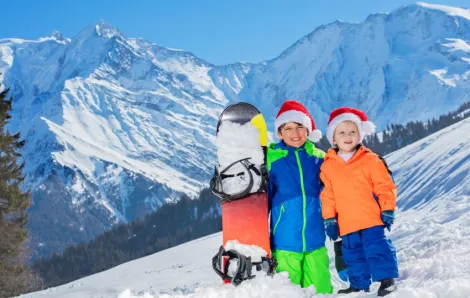 Two happy boys in Santa hats stand with snowboard over the mountains on background on Christmas Day