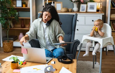Mom on the phone, looking at a computer and holding a tablet doing a lot of things at once. Baby in the background in a highchair watching. 