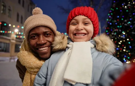 A father and daughter dressed for the cold stand in front of a decorated tree.