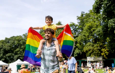 A young boy holds a Pride rainbow flag while on his father's shoulders to celebrate Pride.