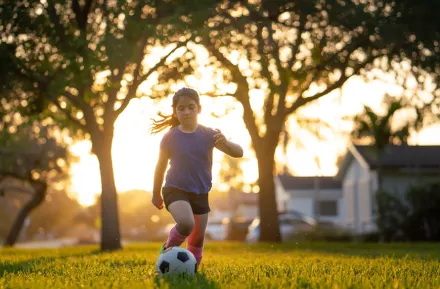 Young girl kicking a soccer ball in a field with trees behind her