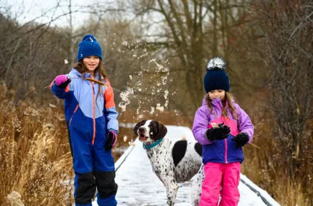 Two girls and their dog on a nature hike in winter. They're wearing winter clothes and there's a dusting of snow on the ground