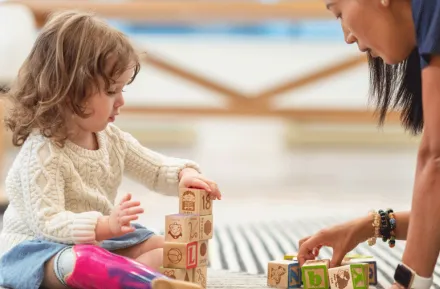 a young girl plays with wooden blocks along with an adult on a rug, safe toys