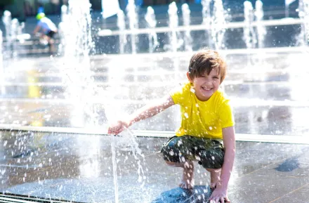 A young boy in a yellow shirt crouches down at a splash pad and enjoys the water.