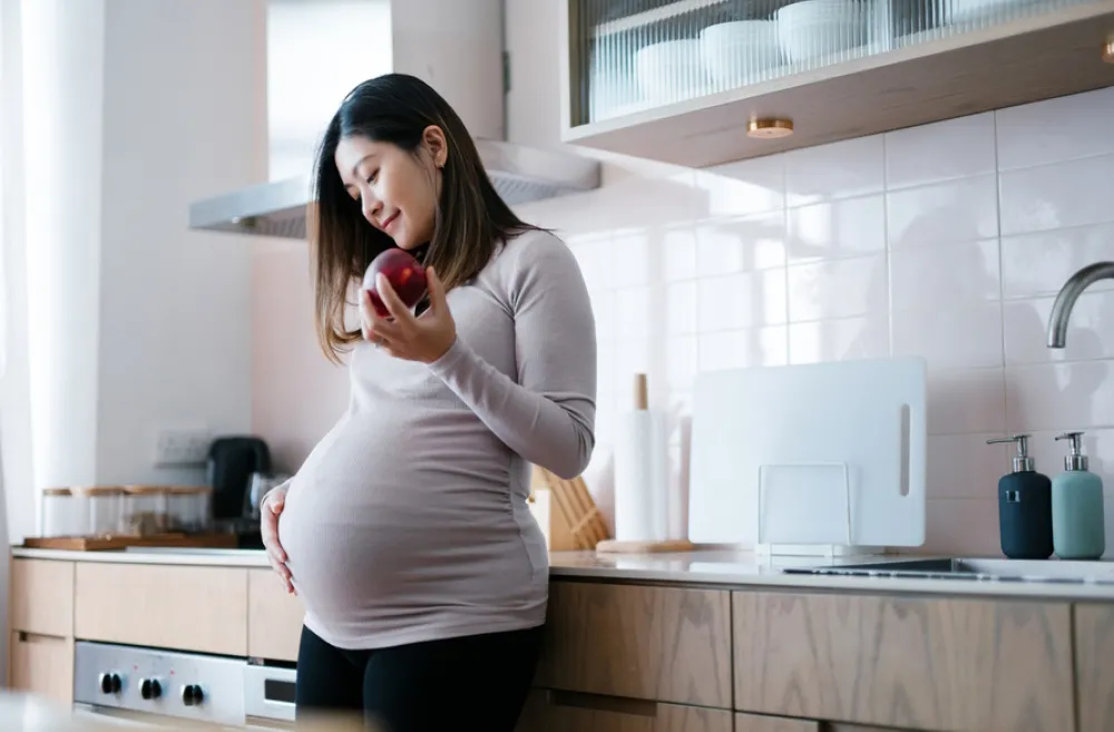 Pregnant woman standing in a kitchen holding an apple and looking down at her belly 