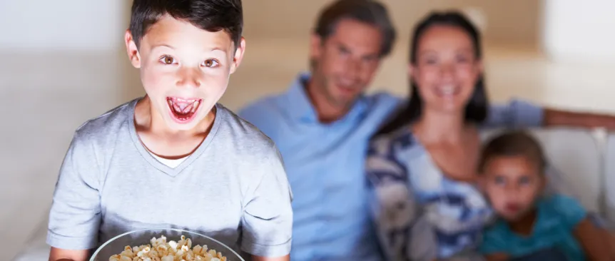 boy holding a bowl of popcorn with a plastic spider in it watching a kids Halloween movie with his parents in the background