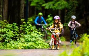 A young girl rides a bike on a bike path with her older sibling and parent following behind. They are wearing helmets and the paved path is lined with green plants indicating summer.