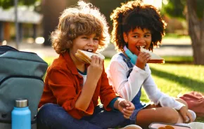 Two kids eating lunch together outside on a sunny day at school 