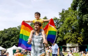 A young boy holds a Pride rainbow flag while on his father's shoulders to celebrate Pride.