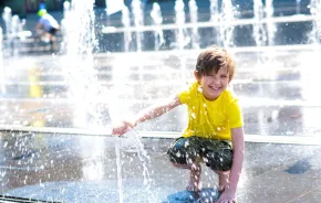 A young boy in a yellow shirt crouches down at a splash pad and enjoys the water.