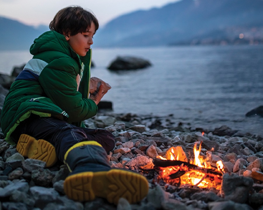A young camper sits beside a fire on the shores of a lake