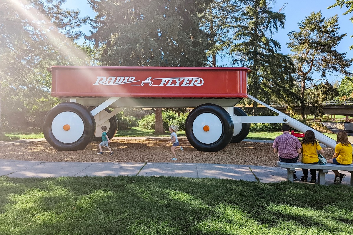 Young kids run in front of a giant red Radio Flyer wagon sculpture in Spokane's Riverfront Park. Kids can slide down the handle.