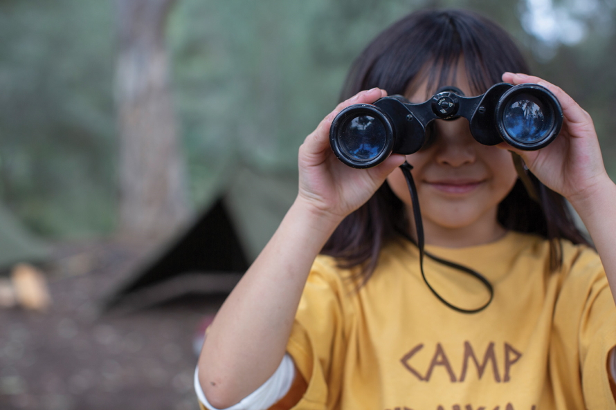 Young girl standing in front of a tent looking through binoculars 
