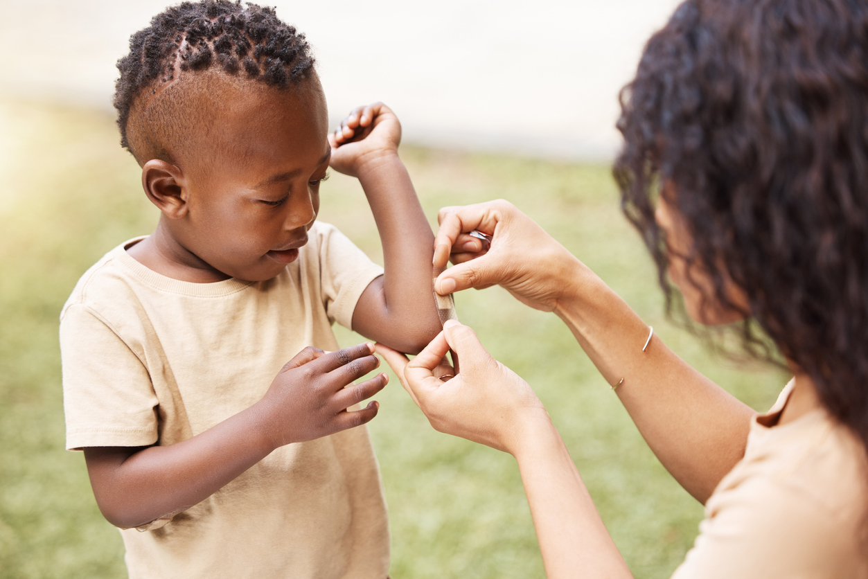 A young child gets a bandaid gently placed on his elbow by a caregiver.