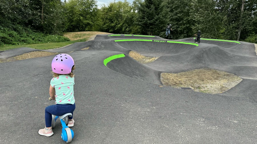 young girl on a balance bike at Jennings Park, a Seattle-area pump track, watching other riders on the track