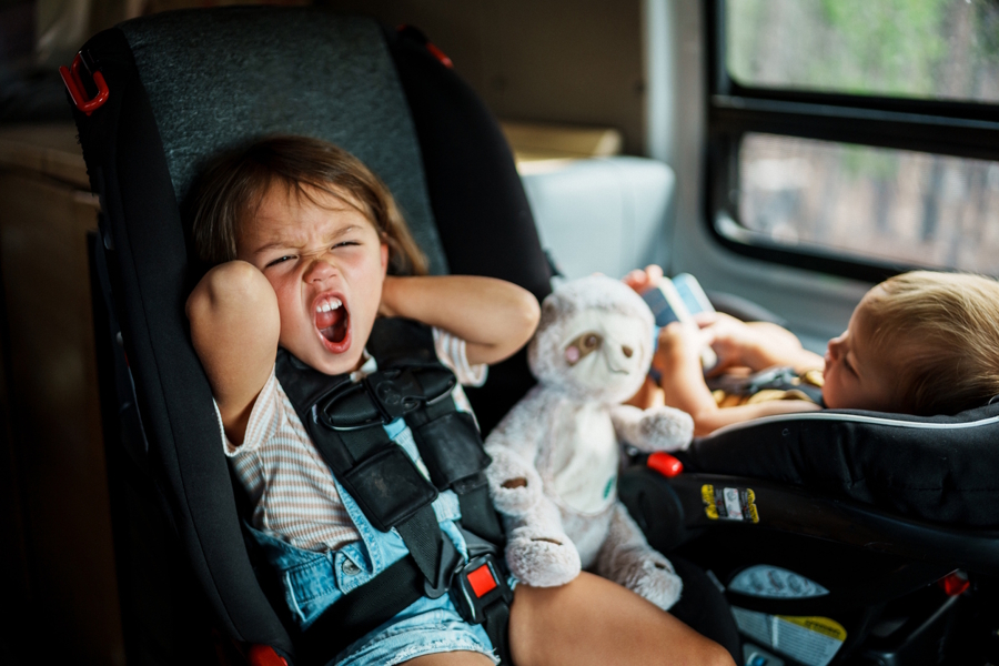 Upset toddler in a car sitting next to a baby on a family road trip