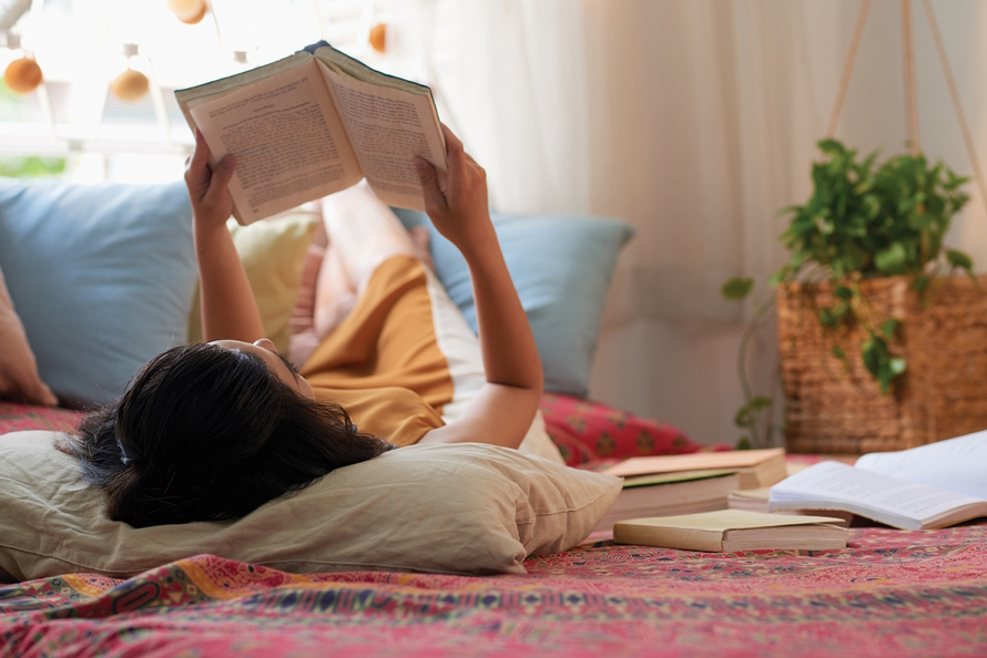 Woman laying on her back on a bed holding up a book and reading