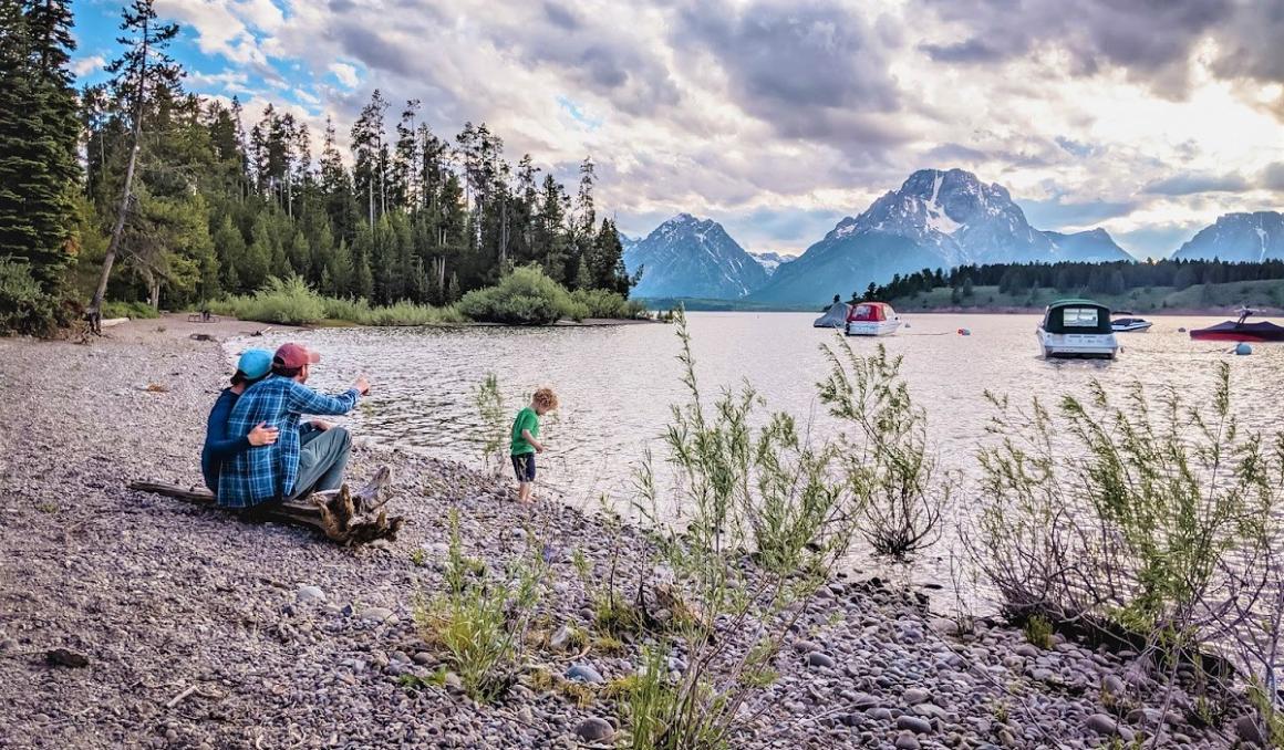 Family sitting on the bank of a river with beautiful mountain scenery in the background camping on a camper van road trip