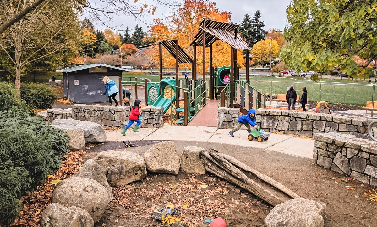 Kids play at updated Lakewood Playground in South Seattle. There's a sand pit in the foreground and a new play structure in the background and fall foliage viewable as well