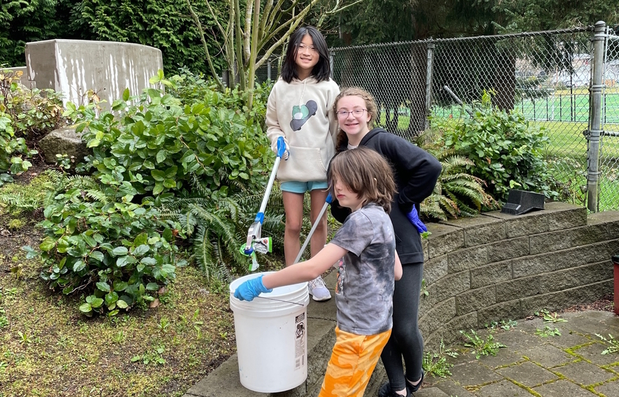 Three elementary kids holding buckets cleaning up an outdoor area