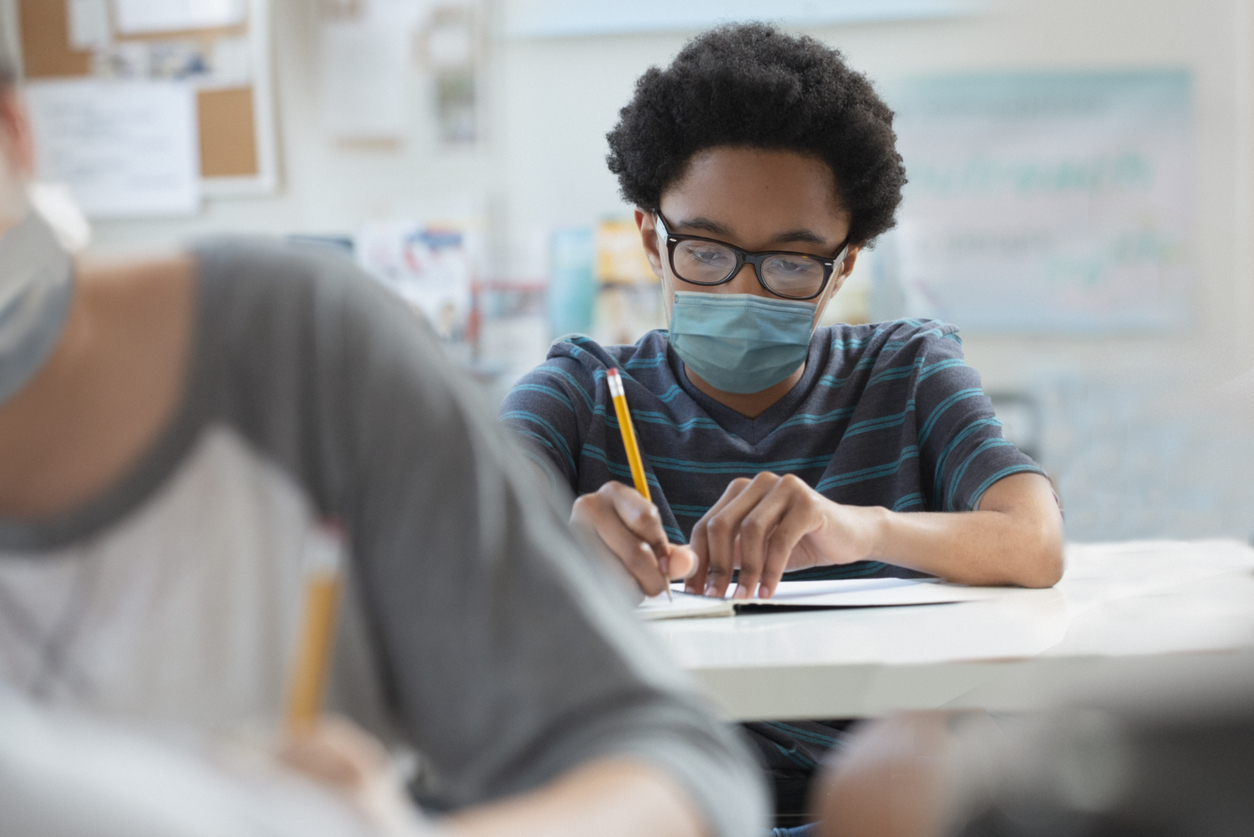 boy wearing glasses and a mask writing with a pencil at his desk in a school classroom