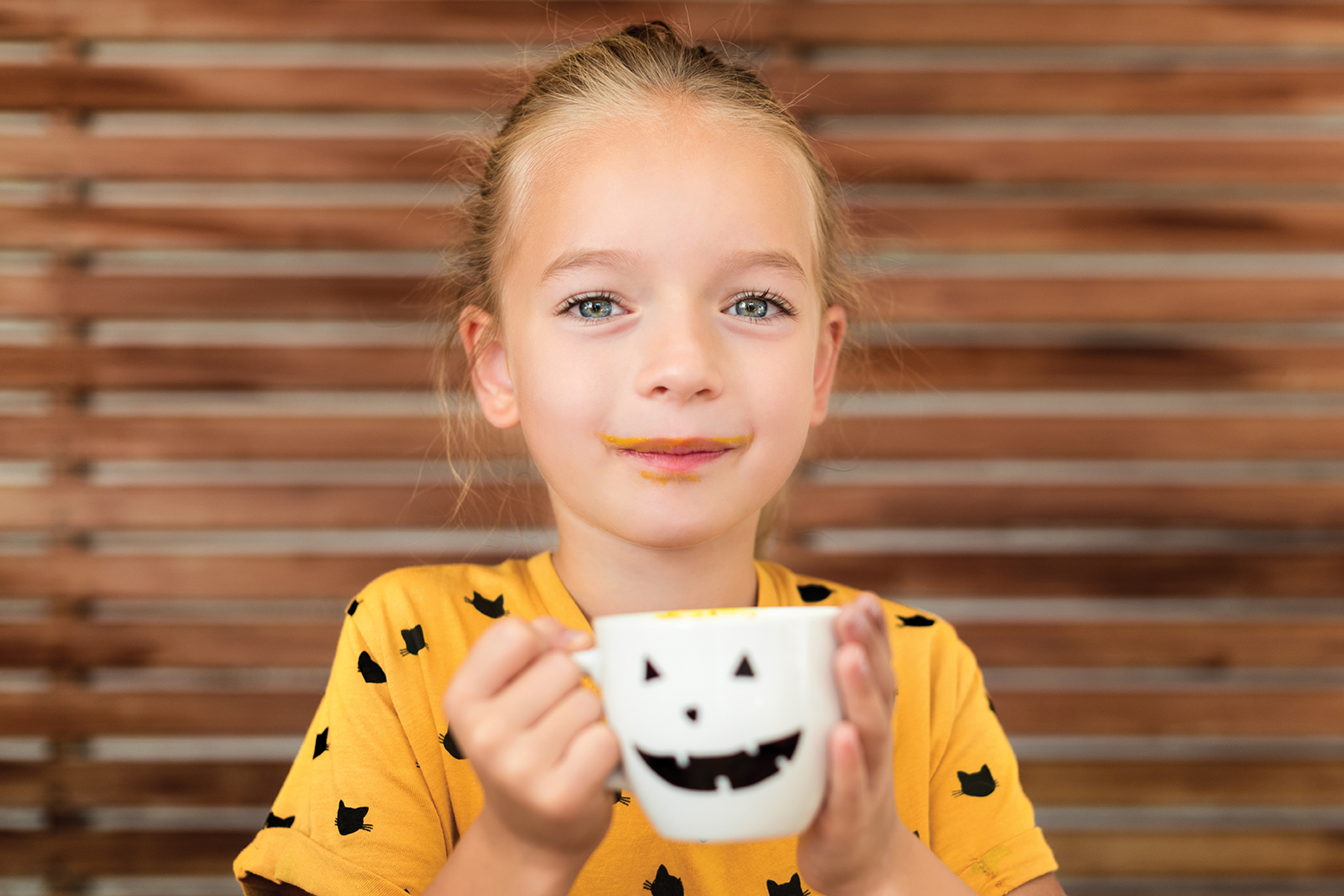 girl in an orange halloween shirt drinking out of a white jack o'lantern mug