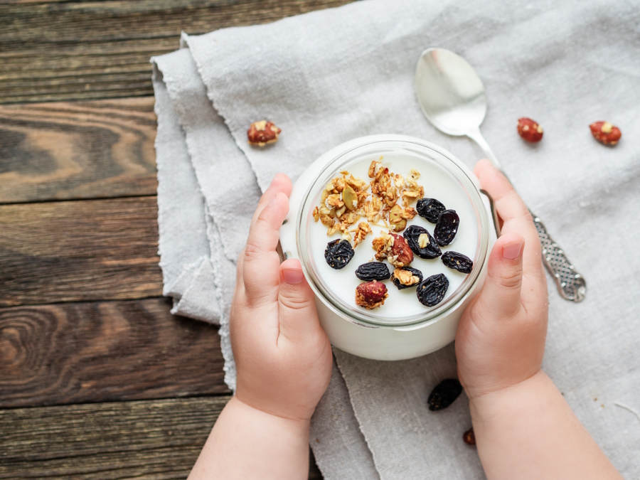 Toddler hands around a glass jar holding yogurt topped with raisins and nuts