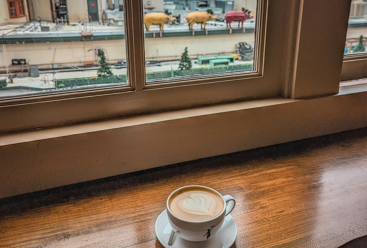 View of a cafe latte at Pike Place Market shop Storyville Coffee with a view of brass pigs through the window