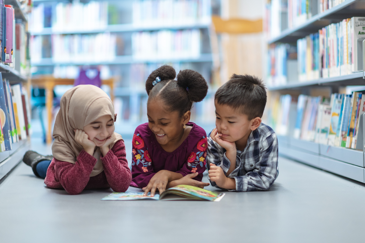 Three kids lying on their stomachs in a library looking at a book together