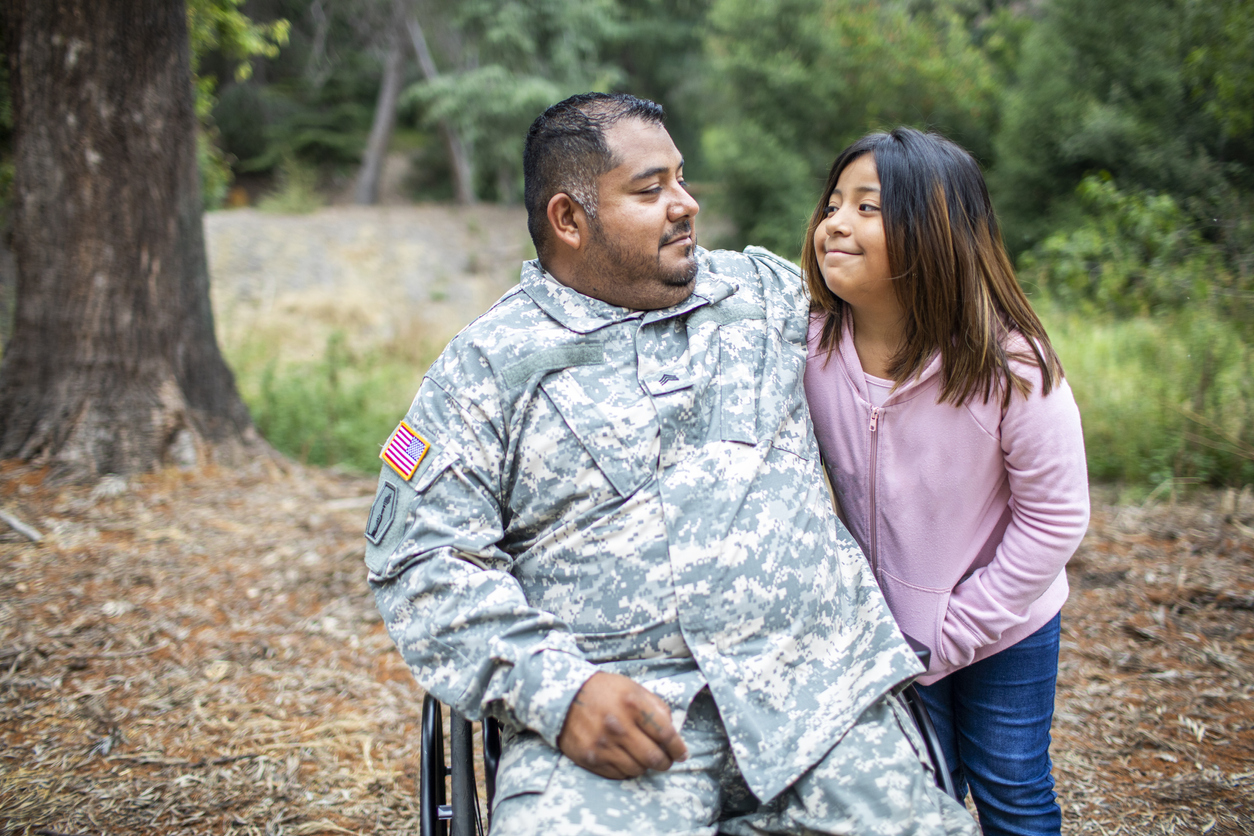 Veteran dad in army fatigues and sitting in wheelchair smiles at and hugs daughter of about age 11. she is smiling at him, wearing a pink sweatshirt