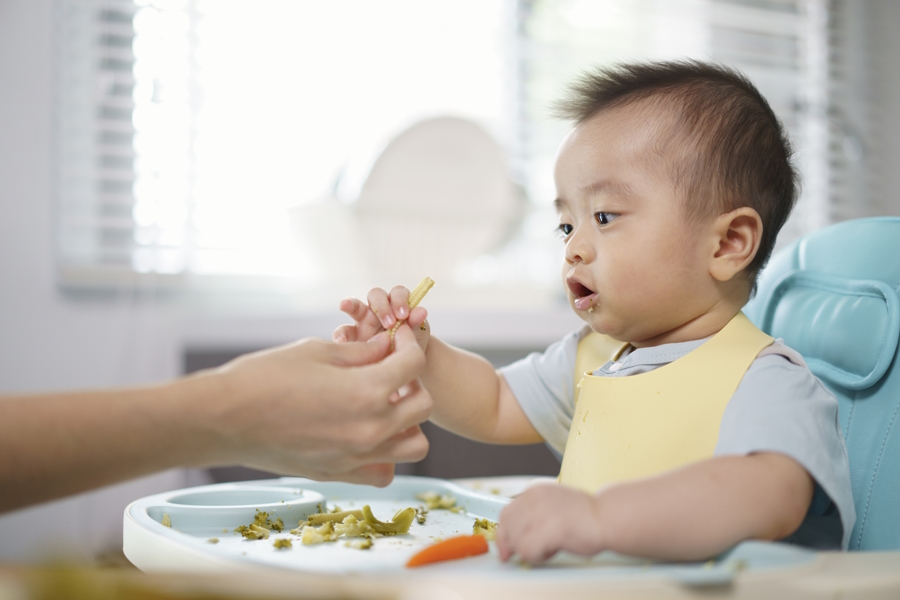 Mom handing baby corn to an infant in a high chair as a way to wean to solid foods