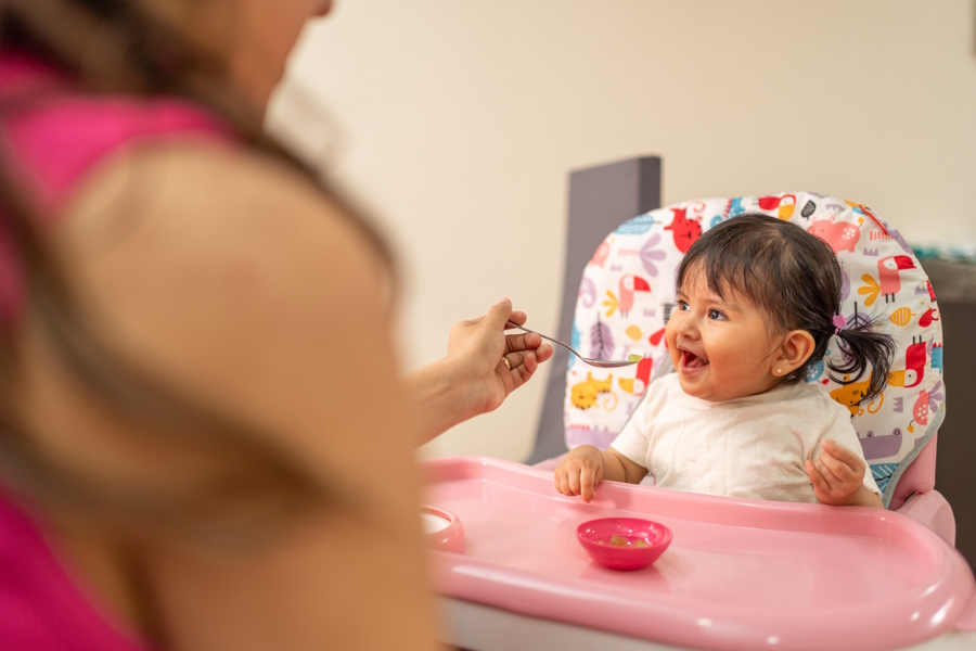 Cute baby smiling and sitting in a highchair while woman feeds her with a spoon, ways to wean to solid foods