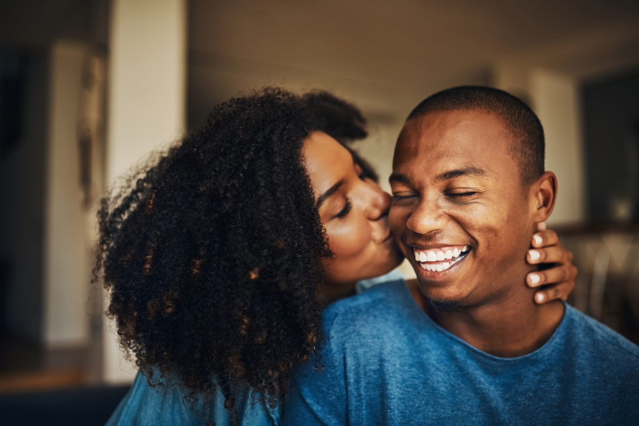 wife hugging her husband from behind kissing his cheek while he smiles