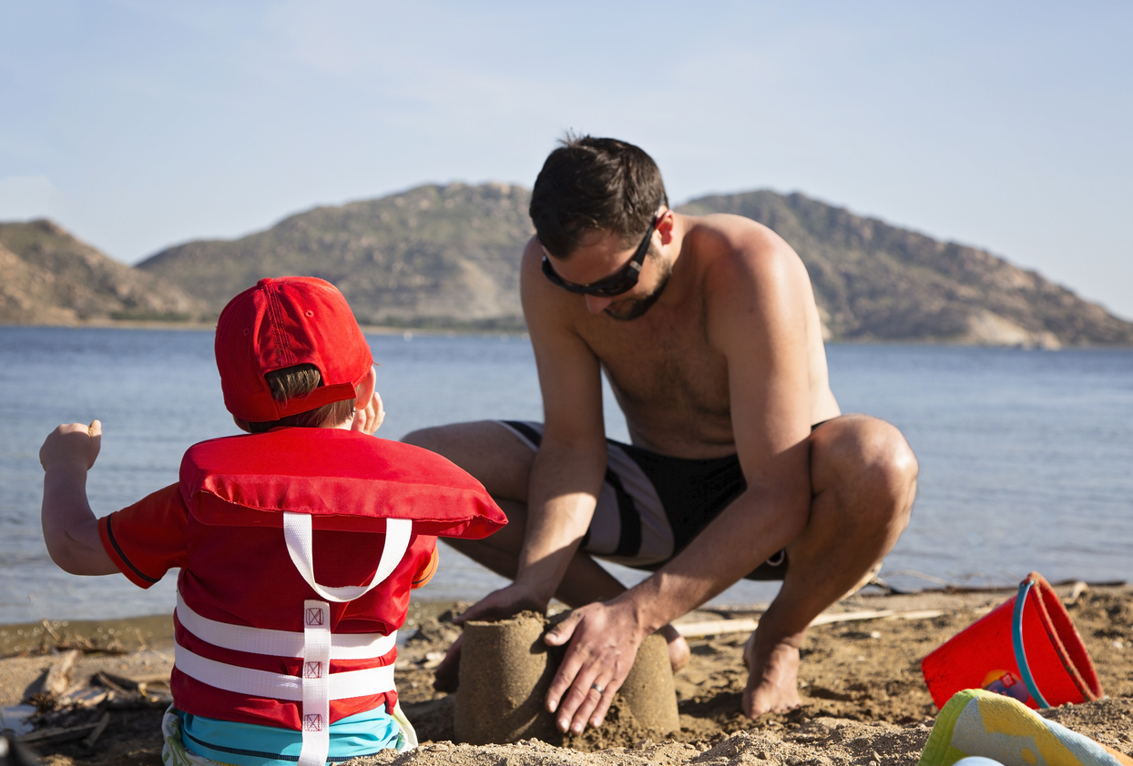 Child wearing a life jacket building in sand with dad