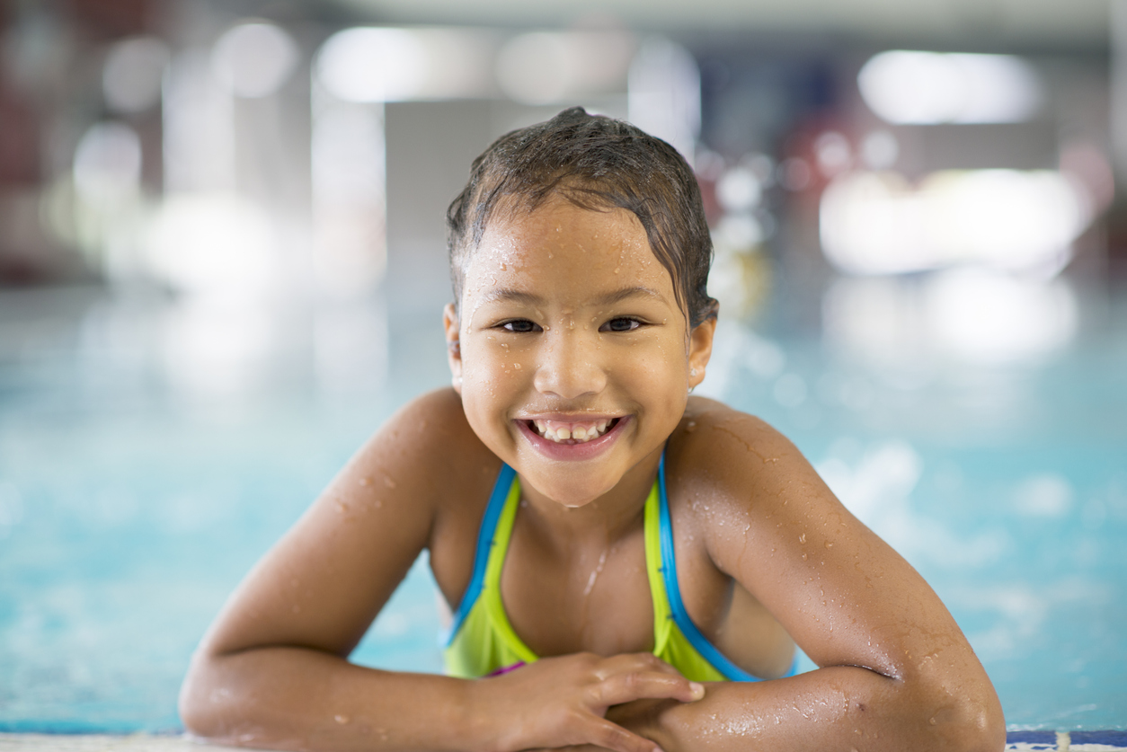 Elementary-age girl smiling, wearing swimsuit, with arms resting on the edge of an indoor swimming pool