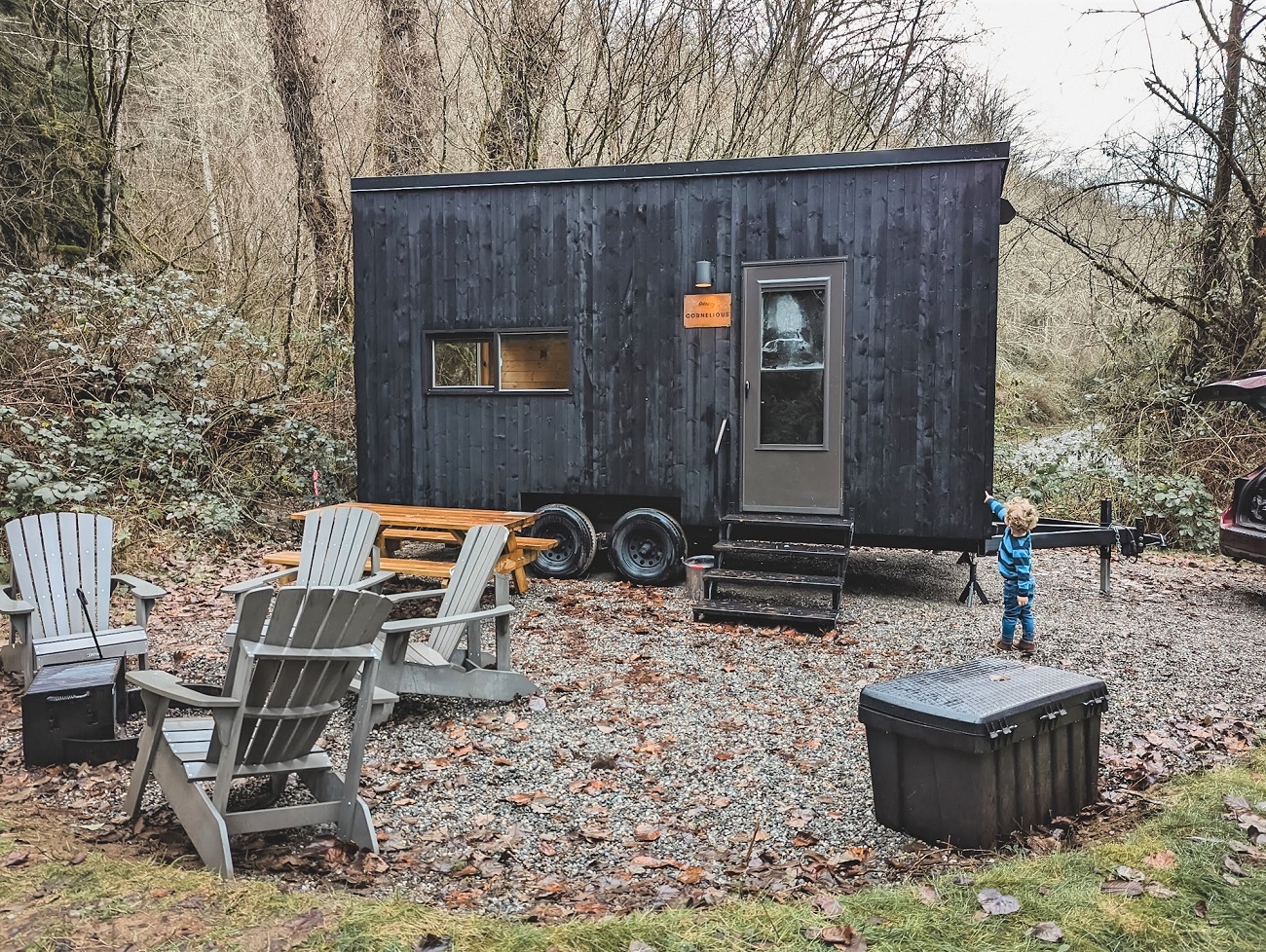 View of Skagit Getaway Cabin a wood-sided trailer set up as a tiny home for rent for yearround family camping. A small boy, chairs and a picnic table are in the foreground