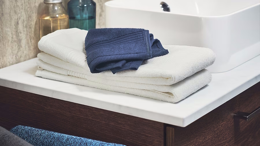 White bath towels and a blue hand towel sitting on the side of the sink hosting essentials from ikea