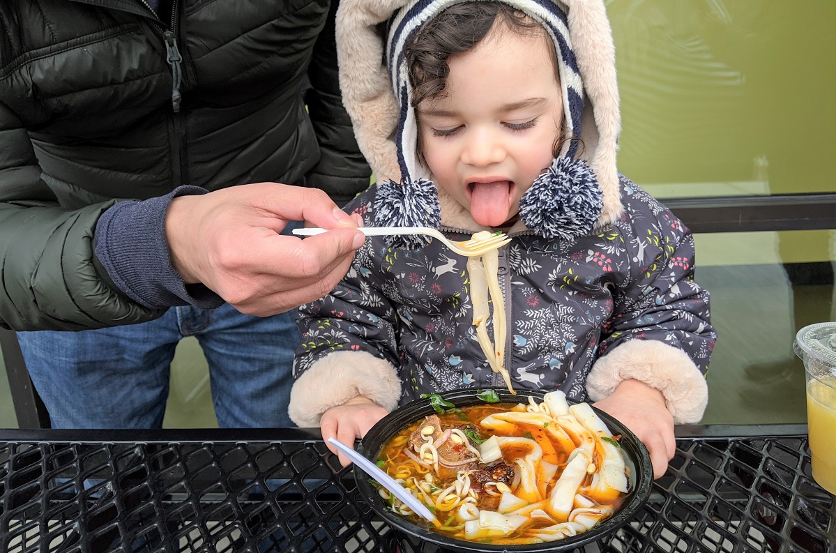 Young girl in warm jacket sitting outside eating, with adult help, a bowl of beef and noodle soup from Theary Spice Bridge Seattle Tukwila